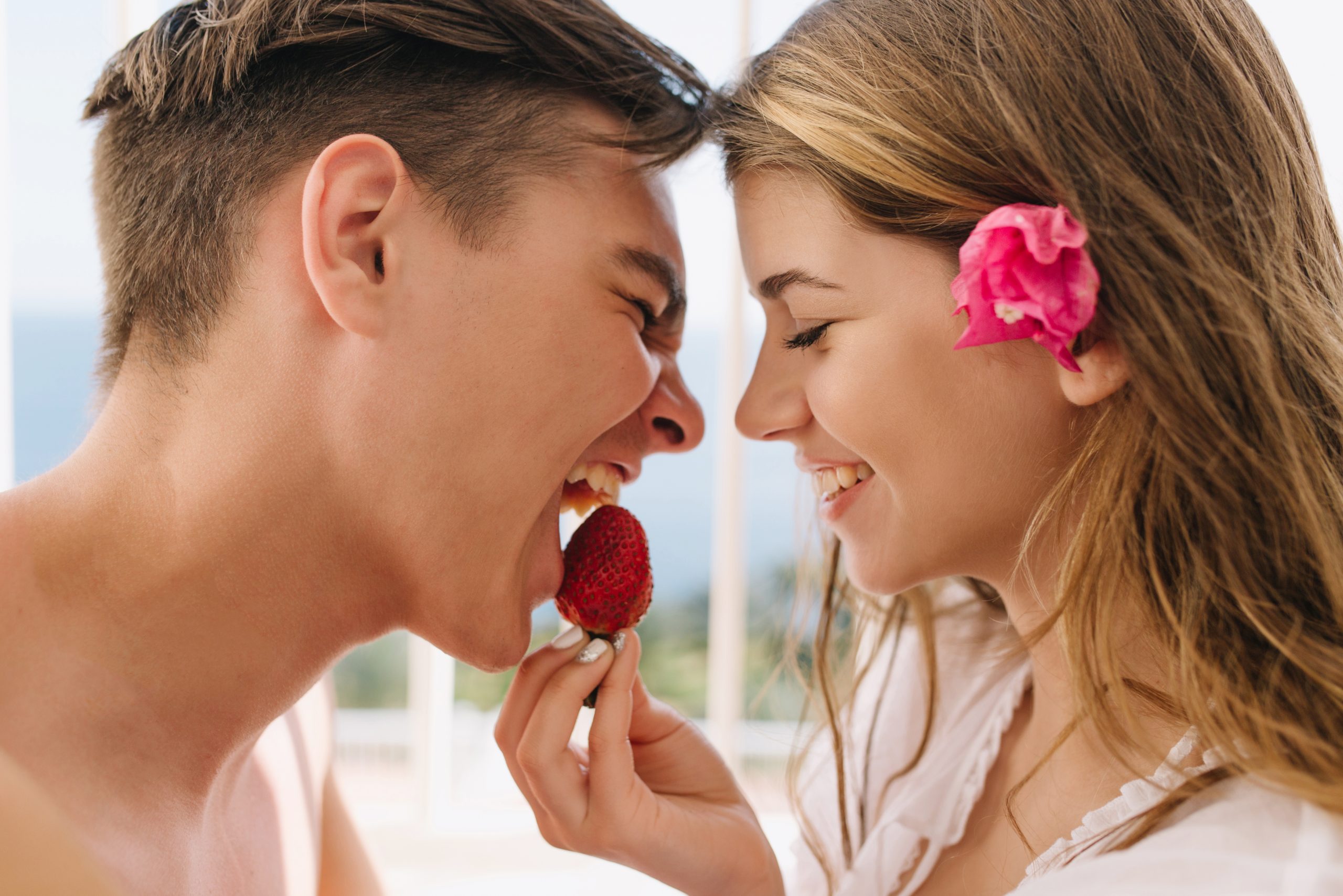 Close-up portrait of loving young couple posing with eyes closed, while eating strawberry on blur background. Adorable blonde girl with pink flower in hair feeds her boyfriend with tasty berries..